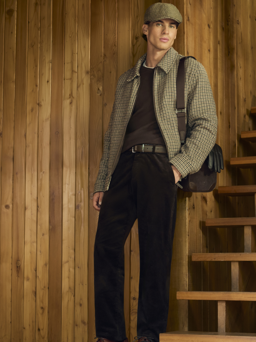 Muž v elegantnom saku s jemným vzorom kohútej stopy, tmavých nohaviciach a klasickej plochej čiapke, stojaci na pozadí drevených schodov.